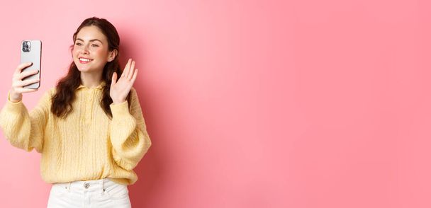 Портрет дружньої дівчини, що розмовляє в додатку для відео чату, махає рукою на камеру смартфона, веде мобільну розмову, стоїть на рожевому фоні
. - Фото, зображення