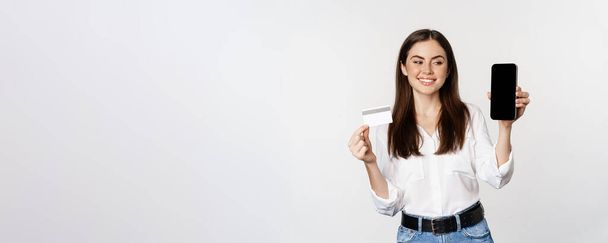 Portrait of female model showing credit card with smartphone screen, recommending application, standing over white background. - Fotó, kép