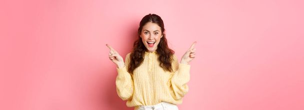 Image of cheerful good-looking female model pointing fingers aside, showing two choices sidewas, left and right logo, standing over pink background. - Fotó, kép