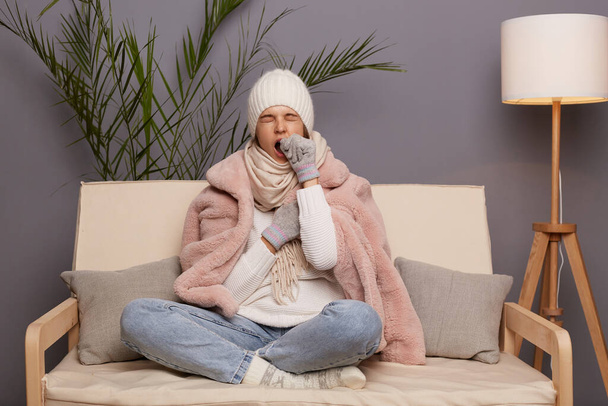 Full length portrait of sleepy woman posing in cold room sitting on the cough wearing warm hat, mittens and cap, needs to have nap, yawning, covering mouth with hand. - 写真・画像