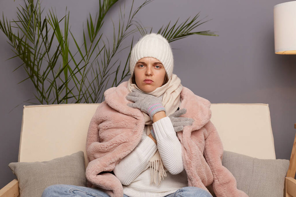 Indoor shot of sad upset woman in cold room sitting on the sofa wearing warm hat, mittens and cap, trying to get warm, looking at camera with sadness and sorrow. - Fotoğraf, Görsel