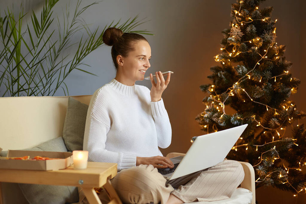 Christmas greetings online. Side view of smiling woman in a white sweater uses a laptop for online work and recording voice messages to friends with congratulating. Happy new year. - Foto, Bild