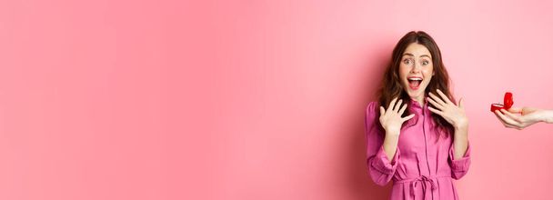 Портрет здивованої дівчини крик збудження, як рука показує її заручини кільце, отримати пропозицію шлюбу, подивитися на камеру здивовано, стоячи над рожевим тлом
. - Фото, зображення