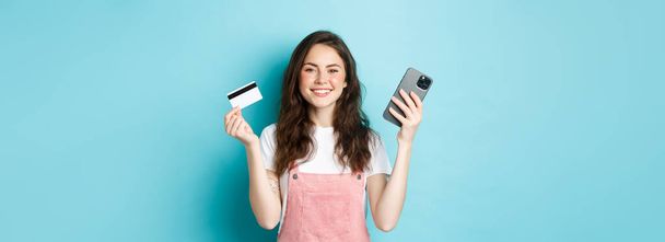 プラスチッククレジットカードやスマートフォンを見せるかわいい女の子を笑顔にし、モバイルアプリ、オンラインショップ、または注文の支払いで店内で何かを注文し、青い背景を背景に立って. - 写真・画像