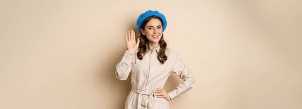 Image of beautiful friendly woman in outerwear, saying hello, waving hand and smiling, greeting you, standing over beige background. - Fotografie, Obrázek