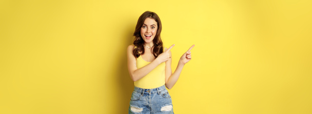 Portrait of stylish summer girl, brunette woman showing way, pointing fingers left, advertising, standing over yellow background. Copy space - Fotoğraf, Görsel