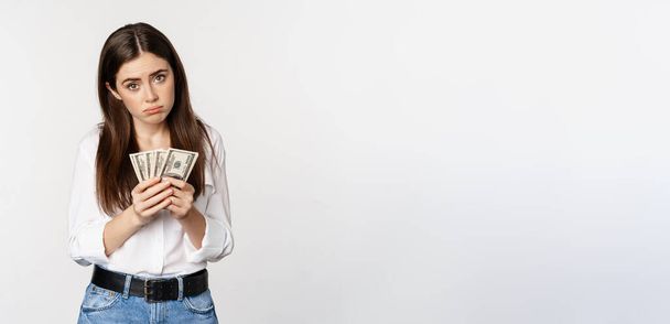 Гломія засмучена дівчина рахує гроші доларів і сумує, стоячи в повсякденному одязі на білому тлі
. - Фото, зображення