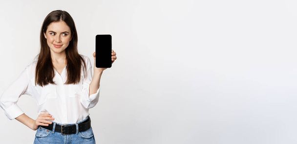 Smiling woman showing mobile phone screen, looking cunning, standing over white background. Copy space - Foto, afbeelding