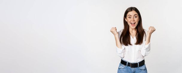 Excited young adult woman reacting to win, surprise news, screaming and cheering, triumphing, achieve goal and celebrating, standing over white background. - Photo, Image