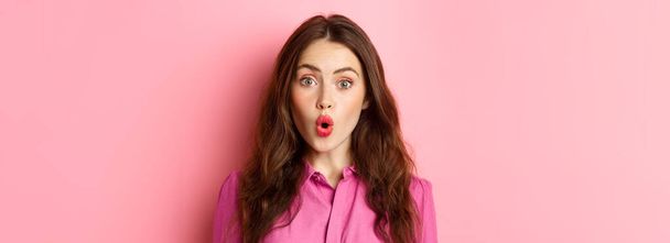 Close up of intrigued young woman gasping, saying wow and looking impressed at camera, standing over pink background. Copy space - Foto, imagen