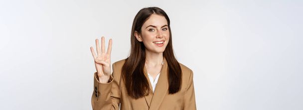 Portret van een zakenvrouw, verkoopster met nummer vier vingers en lachende, staande in pak over een witte achtergrond. - Foto, afbeelding