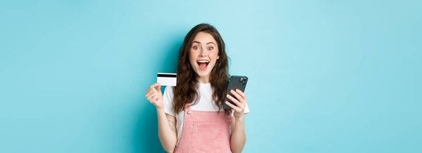 Überraschte und glückliche junge Frau zeigt Plastikkreditkarte und Smartphone, blickt erstaunt in die Kamera, kauft online mit Frühjahrsrabatten ein, blauer Hintergrund. - Foto, Bild