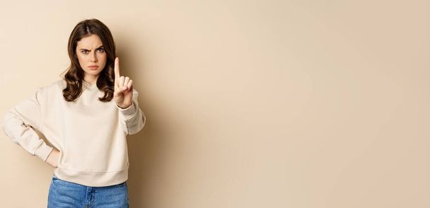 Serious woman show one finger, stop disapproval sign, forbid smth, say no, prohibit, standing over beige background. - 写真・画像
