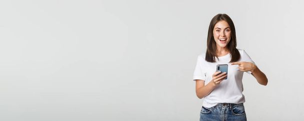 Excited and pleased brunette girl pointing finger at smartphone screen. - Foto, immagini