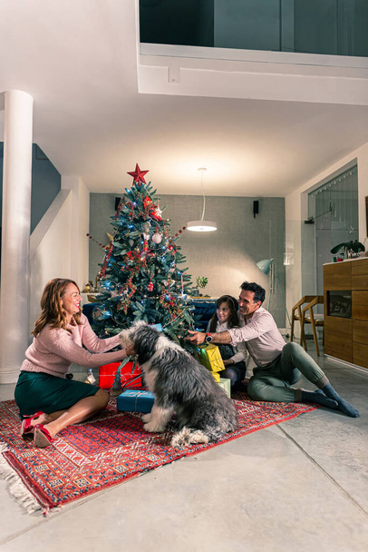 Egy anya és apa ül a lányukkal a padlón a karácsonyfa mellett, és karácsonyi ajándékokat cserélnek. A nappali díszített szellemében a közelgő ünnepek. - Fotó, kép