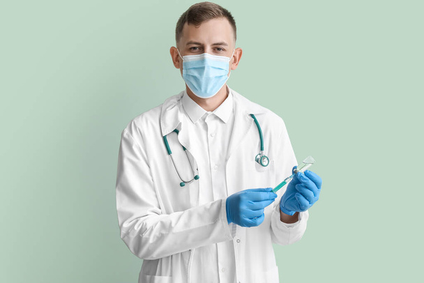 Médico varón en máscara médica con jeringa y ampolla sobre fondo verde - Foto, Imagen