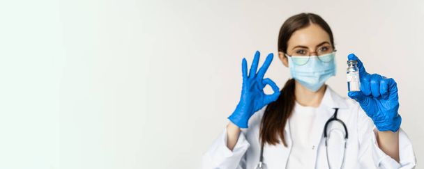 Portrait en gros plan d'une jeune femme médecin professionnelle, médecin portant un masque médical et des lunettes, montrant le vaccin et l'approuvant, recommandant de se faire vacciner contre la covide-19. - Photo, image