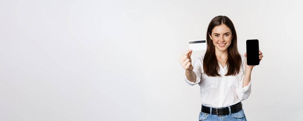 Portrait of female model showing credit card with smartphone screen, recommending application, standing over white background. - Photo, Image