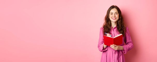 Fröhliches Mädchen lächelt und schreibt Notizen, hält ihren Planer, Plan oder Einkaufsliste in der Hand, steht mit Tagebuch vor rosa Hintergrund. - Foto, Bild