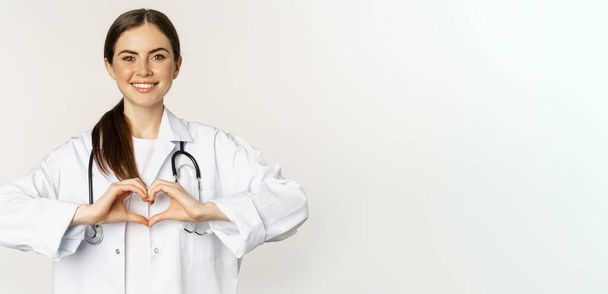 Портрет усміхненого лікаря, жінки-лікаря, що показує знак серця і з обережністю дивиться, кохає своїх пацієнтів, стоїть на білому тлі
. - Фото, зображення