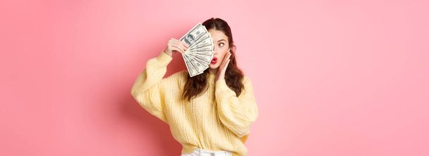 Coquettish young lady pucker lips, making silly face, covering half of it with money dollars, standing over pink background. Copy space - Fotoğraf, Görsel