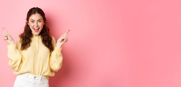 Image d'un beau modèle féminin joyeux pointant les doigts de côté, montrant deux choix latéraux, logo gauche et droit, debout sur fond rose. - Photo, image