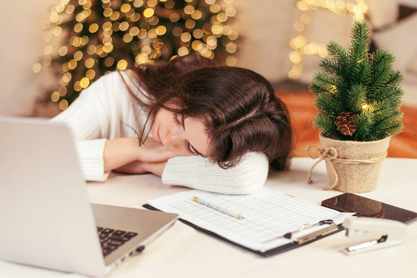 Молодая женщина в белом свитере спит за столом с ноутбуком. Домашнее исследование во время коронавирусного карантина на фоне рождественской елки. Выборочный фокус. Рождественские каникулы - Фото, изображение