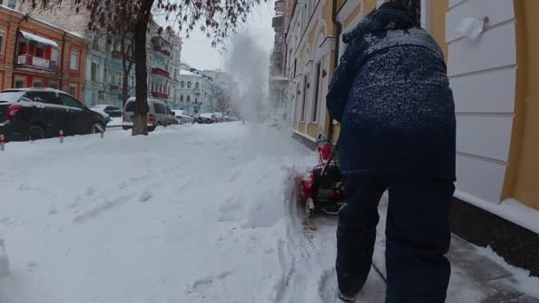  Ένας εργάτης με πράσινη φόρμα αφαιρεί το χιόνι με ένα κόκκινο φυσητήρα στο φόντο ενός έλατου, μιας χιονοστιβάδας και ενός σπιτιού. αργή κίνηση - Πλάνα, βίντεο