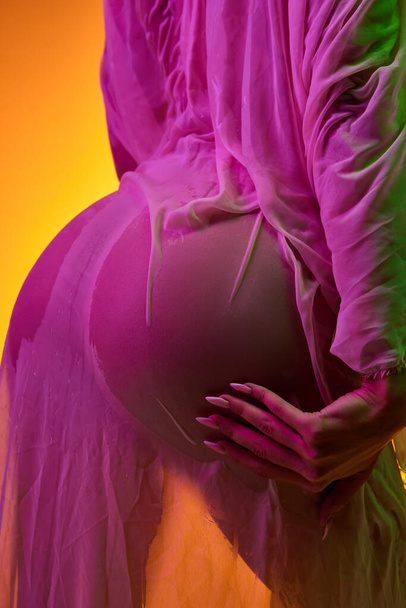 Cropped image of tender female body, buttocks in transparent wet fabric isolated over orange background in pink neon light. Body art, aesthetics. Concept of beauty, sensuality, femininity, skincare. - Photo, image