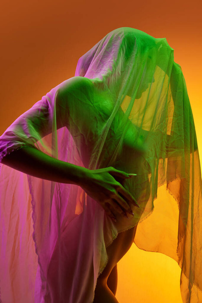 Silhouette di tenera donna rivestita in tessuto trasparente isolato su sfondo arancione in luce verde al neon. Body art, estetica. Concetto di bellezza, sensualità, femminilità, cura della pelle, amore di sé. - Foto, immagini