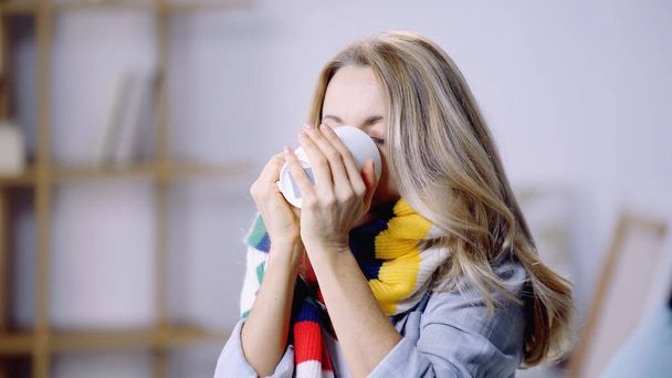 sick blonde woman in colorful scarf holding cup while drinking tea at home   - Fotografie, Obrázek