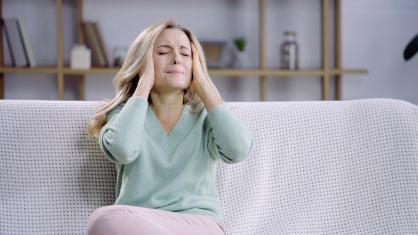 blonde woman with closed eyes having migraine while touching head in living room - Foto, Imagen