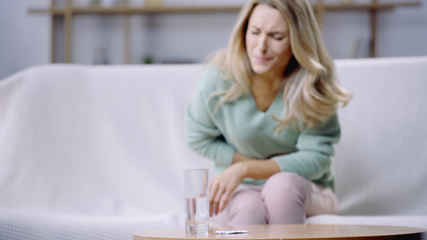 szőke nő szenved menstruációs fájdalom közel pohár víz és üveg pirulák dohányzóasztalon  - Fotó, kép