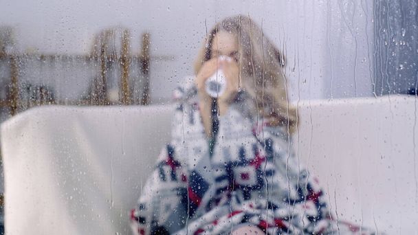 sick woman sitting under colorful blanket and drinking tea behind wet window with rain drops  - Photo, Image