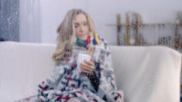 sick woman sitting under colorful blanket and holding cup of tea behind wet window with rain drops  - Фото, изображение