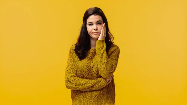 displeased brunette woman in sweater touching cheek isolated on yellow - Foto, Imagem