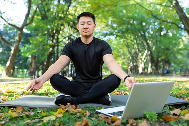 Asian meditating exercise in lotus pose, man resting after fitness training using laptop for online meditation training. - 写真・画像