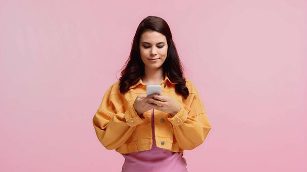 smiling young woman in orange jacket texting on smartphone isolated on pink - 写真・画像