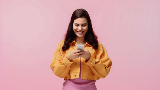 happy young woman in orange denim jacket texting on smartphone isolated on pink - Fotó, kép