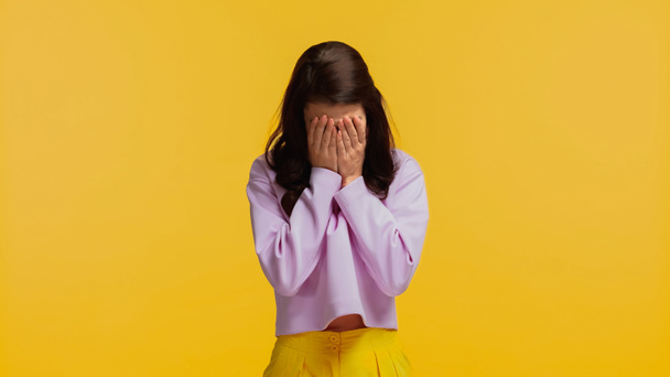 brunette vrouw in paars sweatshirt bedekking gezicht terwijl huilen geïsoleerd op geel - Foto, afbeelding