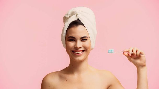 счастливая молодая женщина с голыми плечами и полотенцем на голове держа зубную щетку с зубной пастой изолированы на розовый  - Фото, изображение