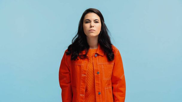 displeased and brunette woman in orange denim jacket looking at camera isolated on blue - Фото, изображение