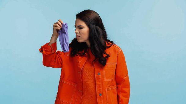 disgusted woman in orange jacket holding stinky socks isolated on blue - Foto, Bild