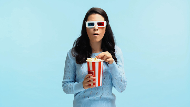geschokt jonge vrouw in 3D bril eten popcorn terwijl het kijken naar film geïsoleerd op blauw - Foto, afbeelding