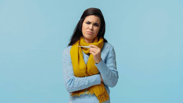 αναστατωμένη και άρρωστη γυναίκα με κασκόλ που κρατά ηλεκτρονικό θερμόμετρο απομονωμένο στο μπλε - Φωτογραφία, εικόνα