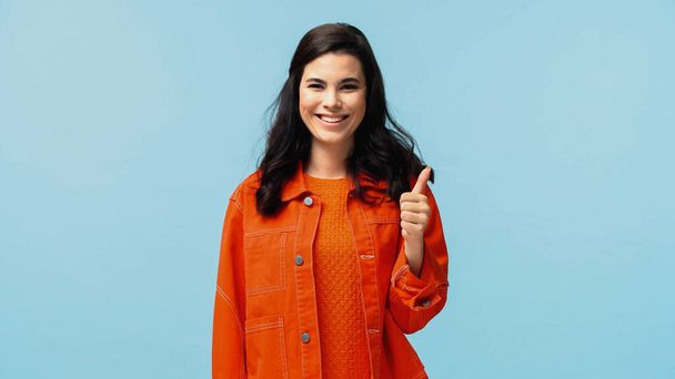 cheerful young woman in orange jacket showing thumb up isolated on blue - Fotografie, Obrázek
