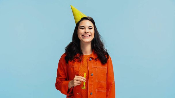 cheerful young woman in orange jacket and party cap holding festive horn while laughing isolated on blue - 写真・画像