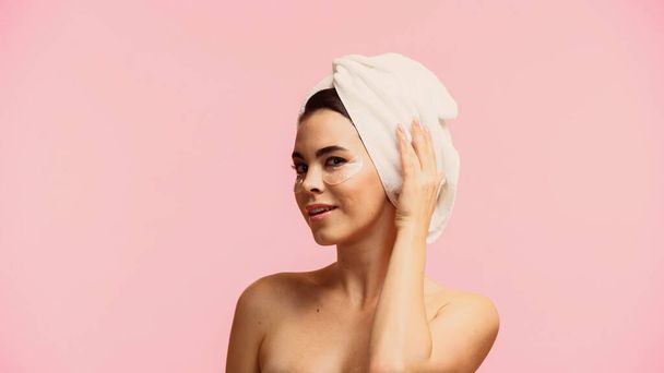 gelukkig jong vrouw met blote schouders en ooglapjes aanraken handdoek op hoofd geïsoleerd op roze  - Foto, afbeelding