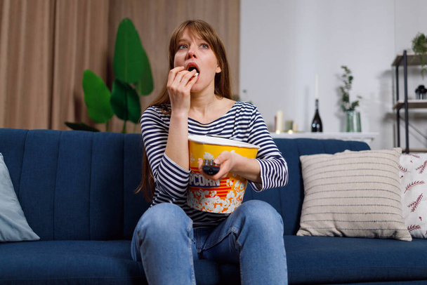 Geconcentreerde jonge vrouw met popcorn emmer kijken tv thuis - Foto, afbeelding