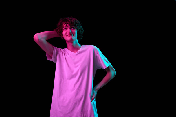 Smiling young man looking up dreamily isolated over dark background in purple neon light. Positive emotions, youth, sales, ad. Model, actor wearing white cotton t-shirt - Photo, image